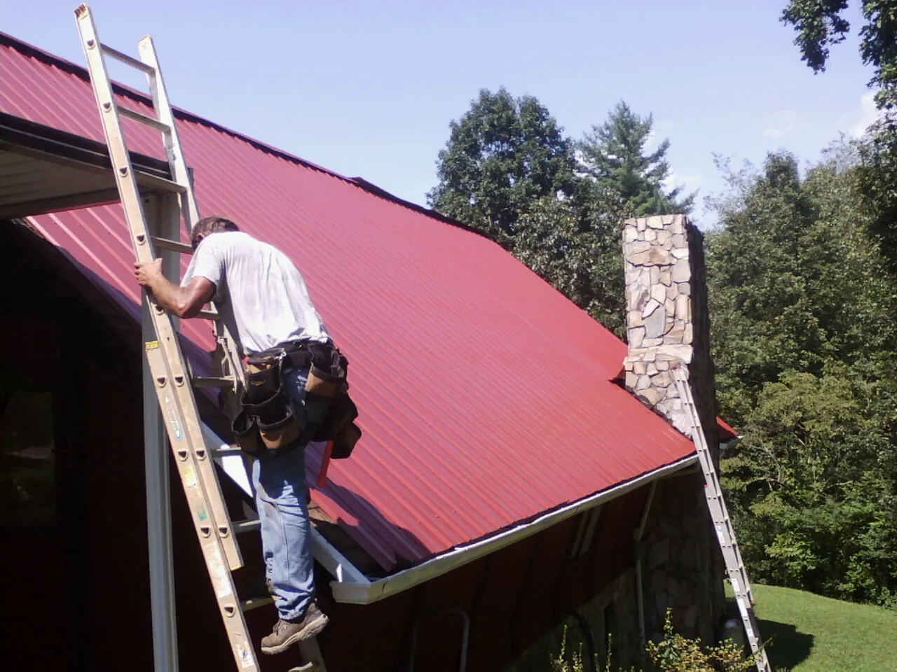Roofer on a ladder beside a red metal roof and stone chimney — Soffit & Fascia Repair by Roofing Brothers in Stamping Ground